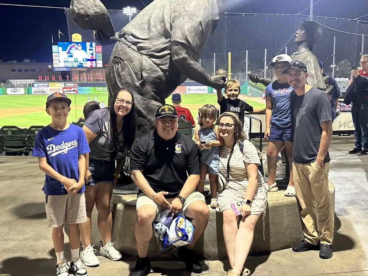 Founders of Operation Hometown Heroes and their families at a minor league ballpark, seated around a bronze statue.